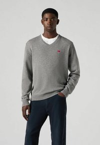 Szary sweter w serek wykonany z miękkiego materiału, z małym czerwonym logo na piersi. Nosi się go na białej koszuli z ciemnymi spodniami.