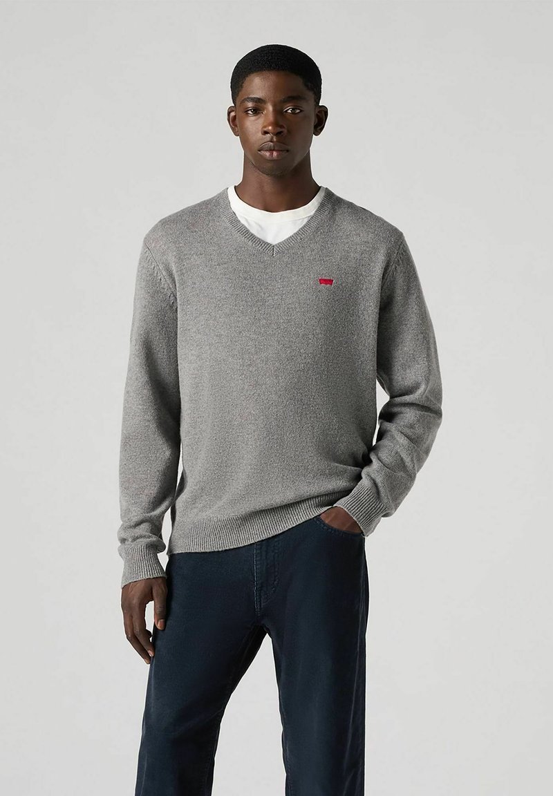 Suéter gris de cuello en v hecho de una tela suave, con un pequeño logo rojo en el pecho. Lleva puesto sobre una camisa blanca con pantalones oscuros.