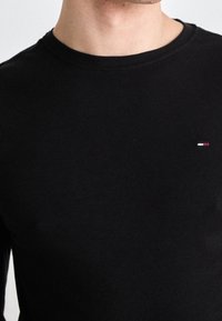 En man klädd i en enkel svart t-shirt med rund halsringning, med en liten rektangulär logotyp i röd, vit och blå färg på vänster bröst.