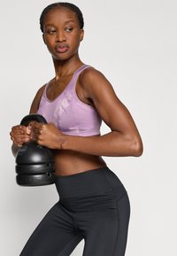 Μωβ αθλητικό σουτιέν με υφή, σετ με μαύρο κολάν με ψηλή μέση. Άτομο κρατάει ένα μαύρο kettlebell.