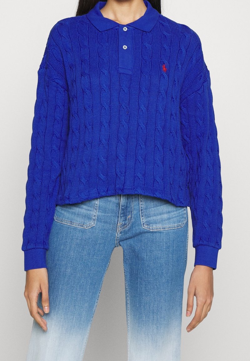 Maglione blu a trecce con colletto polo e tre bottoni, caratterizzato da polsini a costine e un design cropped. Abbinato a jeans azzurri.