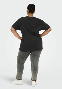 Osoba z włosami związanymi w kok, ubrana w czarny oversize'owy t-shirt, szare dżinsy w stylu acid wash z postrzępionymi brzegami oraz białe sneakersy, stoi z rękami na biodrach.