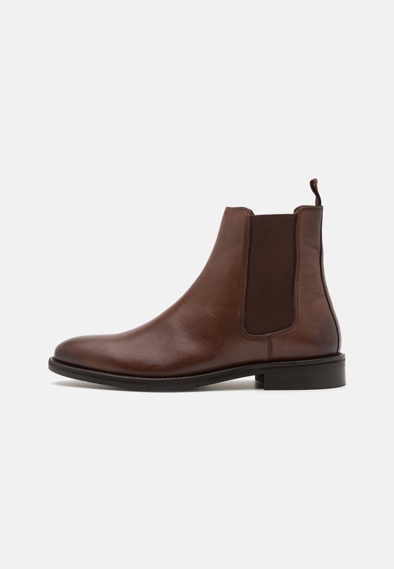 Bottine Chelsea en cuir marron avec panneaux latéraux élastiques, languette au talon et semelle basse noire sur fond blanc.