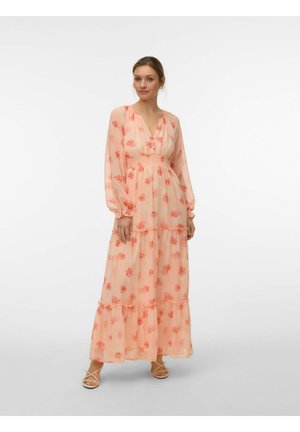 Vrouw draagt een lange, doorschijnende perzikkleurige jurk met rode bloemmotieven, lange mouwen en geplooide lagen, staand met een neutrale uitdrukking tegen een effen achtergrond.