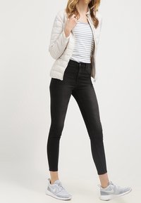 Kvinna som bär en vit quiltad jacka, vit och grå-randig skjorta, svarta tajta jeans och grå Nike-sneakers, stående mot en enfärgad bakgrund.