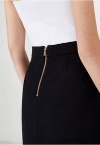 Černá upravená sukně s viditelným zlatým zipem na zadní straně, se hladkou texturou a strukturovaným designem. Na vrchu jednoduchý bílý tílko.