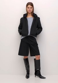 Čierna nepremokavá bunda s kapucňou, sivý sveter, čierne šortky a čierne špicaté čižmy po kolená. Hladké textúry, neformálny strih a monochromatická farebná škála.