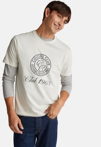 Světle barevné bavlněné tričko s kulatým výstřihem, které má kulatý logo v černém písmu a šedou dlouhou rukávovou vrstvu. Viditelné džíny.