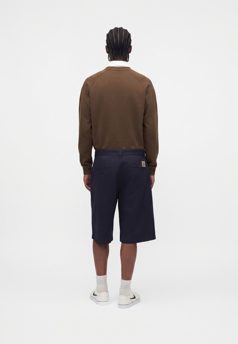 Mand med fletninger står med ryggen mod, iført brun langærmet skjorte, marineblå shorts, hvide sokker og hvide sneakers med sort swoosh.