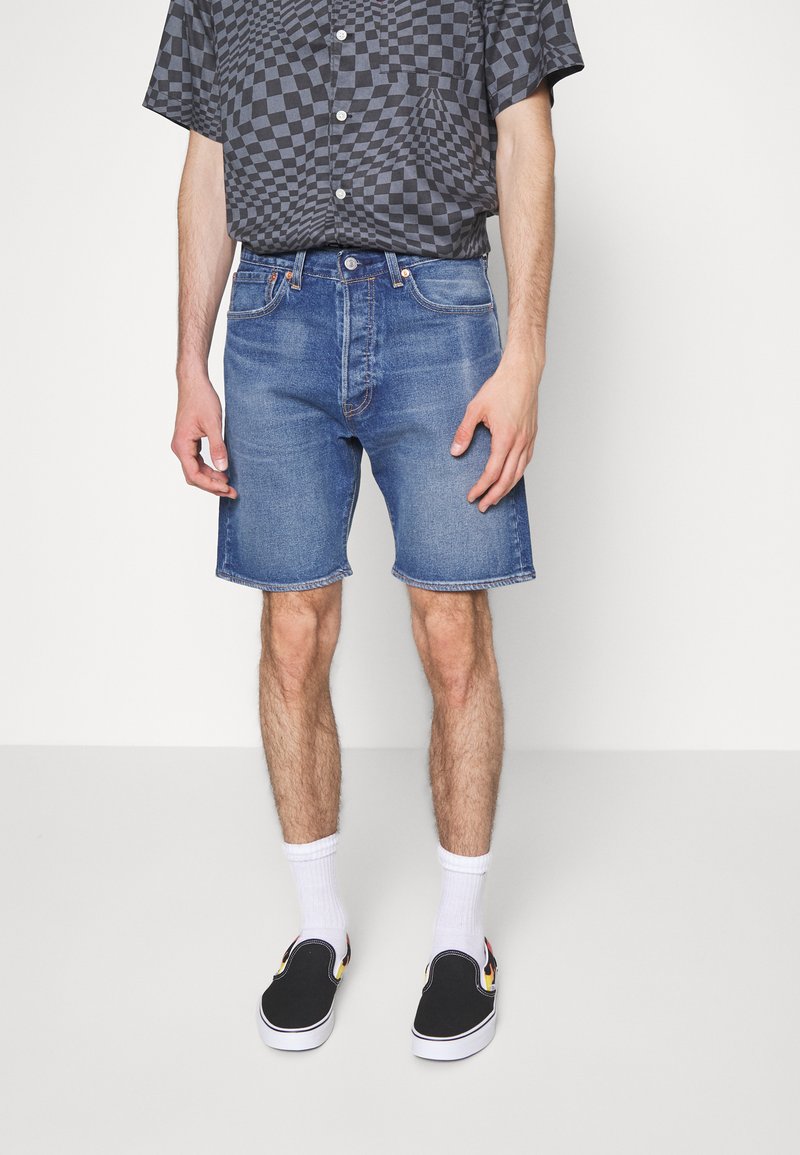 Pantalones cortos de mezclilla en azul medio, con un corte clásico y un dobladillo deshilachado. Combinados con zapatos negros sin cordones y calcetines blancos hasta el tobillo.