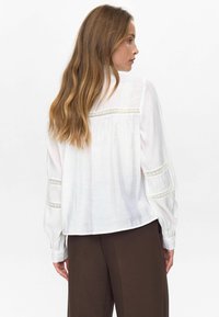 Donna con lunghi capelli mossi che indossa una blusa bianca con dettagli in pizzo sulla schiena e sulle maniche, abbinata a pantaloni marrone scuro, in posizione di profilo.
