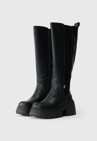 Černé kožené kozačky až ke kolenům s robustní podrážkou, bočním zipem a elastickým panelem pro flexibilitu. Mají texturovaný povrch.