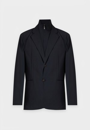 Homme portant un blazer noir, un t-shirt blanc, un jean large bleu clair et des baskets blanches, debout avec une main dans la poche.