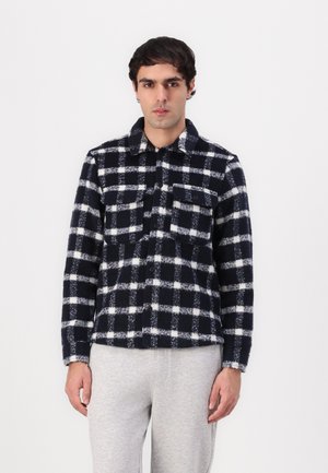 Navy and white checkered flannel shirt with button closure, two chest pockets, and soft, textured fabric. Worn over grey sweatpants.