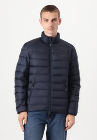 Tumši zila pūķa jaka ar augstu apkakli, priekšpusē rāvējslēdzēju un divām sānu kabatām. Raksturīgas horizontālas šuves un zīmola nosaukums uz krūtīm.