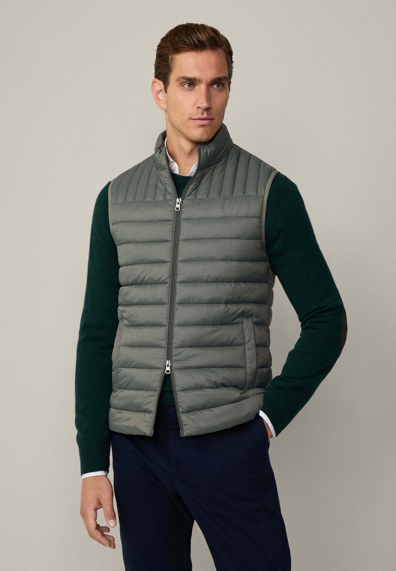 Graue Steppweste mit Frontreißverschluss und Stehkragen, mit horizontalen Steppnähten und seitlichen Taschen, getragen über einem dunkelgrünen Pullover.