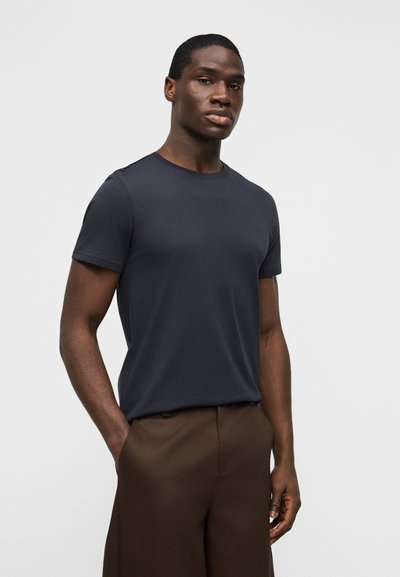 Νέος άνδρας με σκούρο δέρμα, φορώντας εφαρμοστό σκούρο μπλε κοντομάνικο πουκάμισο και καφέ παντελόνι, στέκεται με το ένα χέρι στην τσέπη μπροστά σε λευκό φόντο.