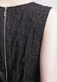 Detailní záběr na černé bezramínkové šaty s květinovou výšivkou a stříbrným zipem na zádech, které jsou oblečeny na světlé ruce.