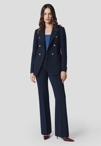 Marynarski żakiet skrojony na miarę z złotymi guzikami, w połączeniu z dopasowanymi spodniami. Garnitur ma dopasowany krój i rozszerzane nogawki, stylizowany z bordowymi szpilkami.
