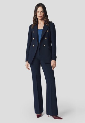 Marynarski żakiet skrojony na miarę z złotymi guzikami, w połączeniu z dopasowanymi spodniami. Garnitur ma dopasowany krój i rozszerzane nogawki, stylizowany z bordowymi szpilkami.