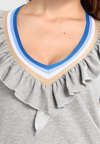 Grå tröja med V-ringning och randigt sicksack i blått, vitt och beige, med en volangdetalj längs halslinningen och ett litet nyckelhålskärning nedanför.