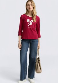 Chemise rouge à manches trois-quarts avec un motif floral blanc et du texte. Associée à un jean en denim bleu et des baskets blanches. Le mannequin tient un sac beige.