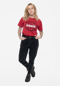 Red cotton t-shirt with "oasis" logo in white, worn with black high-waisted jeans. Black lace-up boots, minimal design, plain background.