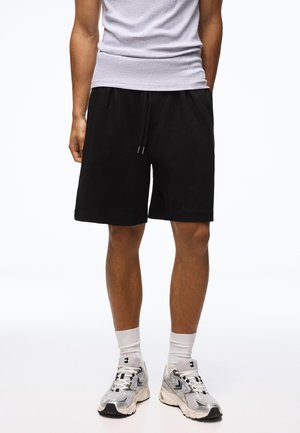 Uomo che indossa pantaloncini neri con coulisse, calzini bianchi, sneakers argento e nere e una maglietta bianca senza maniche, in piedi su sfondo bianco.