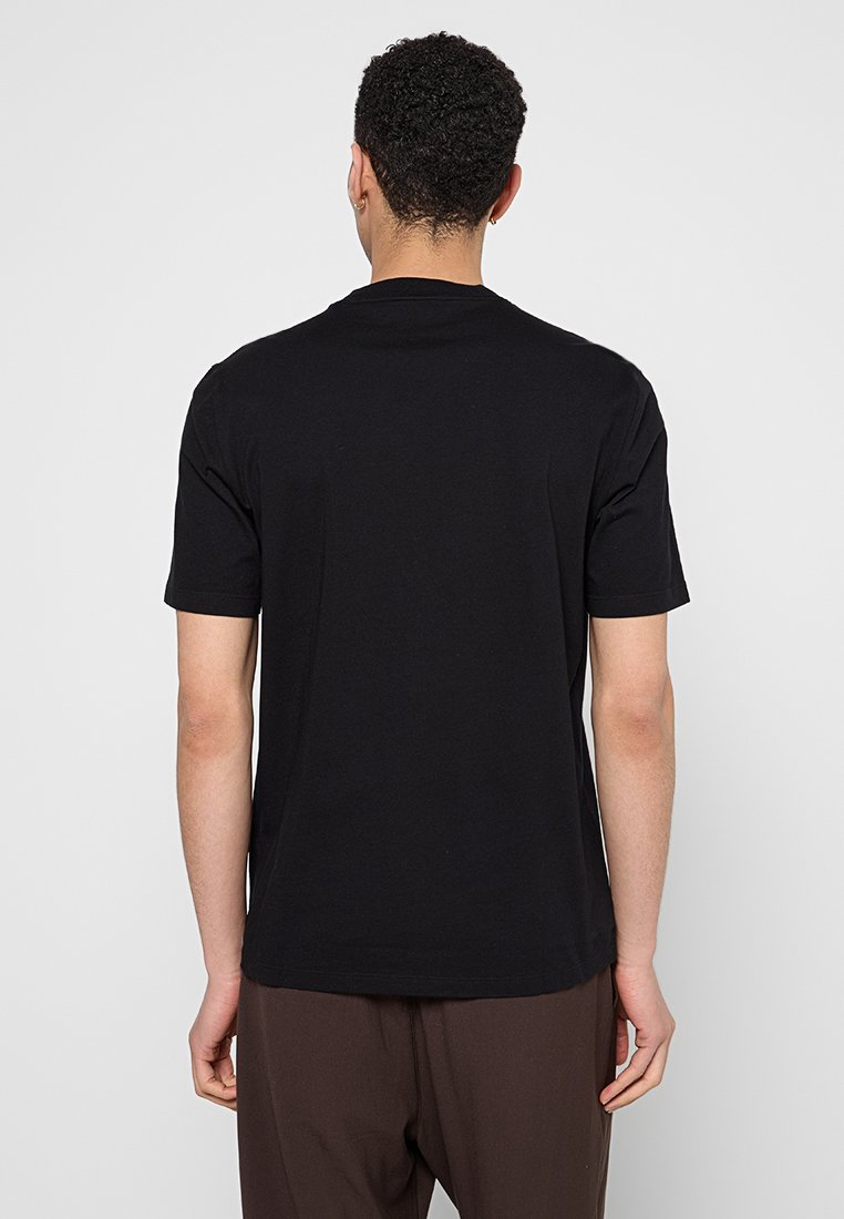 T-shirt noir à manches courtes en coton, avec une coupe décontractée, un col rond et une texture lisse, porté avec un pantalon marron.