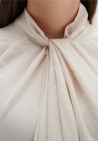 Blusa beige chiaro con un design plissettato e una texture morbida e liscia. Presenta un colletto con fiocco a livello del collo per un tocco di dettaglio.