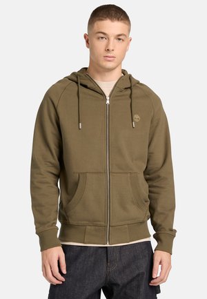 Junger Mann trägt einen olivgrünen Kapuzenpullover mit Reißverschluss, Kordelzügen, Fronttaschen und einem kleinen gestickten Logo auf der Brust, kombiniert mit dunklen Jeans.