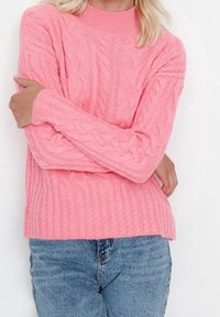 Pull en maille torsadée rose avec un col montant et un ourlet côtelé, associé à un jean en denim bleu. Les manches sont longues avec des détails texturés.
