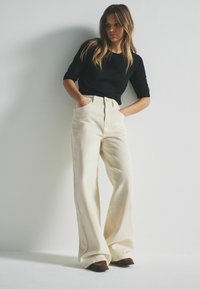 Brede ivoren broek met hoge taille, gemaakt van stevige stof. Voorzien van zijzakken en zichtbare stiksels. Gecoördineerd met een zwart bovenstuk.