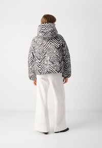 Giacca a vento con motivo zigzag bianco e nero, in tessuto testurizzato, con cappuccio. Abbinata a pantaloni bianchi a gamba larga e calzature nere.
