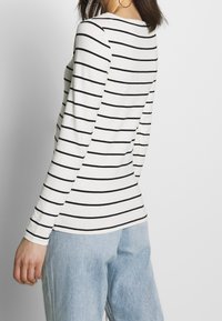 Långärmad topp i vitt med svarta horisontella ränder. Tillverkad av mjukt tyg, har rund halsringning och figursydd siluett.