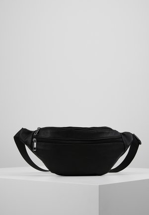 Sac banane en cuir noir avec deux compartiments zippés et une sangle réglable, exposé sur un socle blanc sur fond gris.