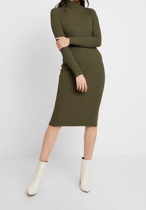 Femme portant une robe midi à col roulé en tricot côtelé vert olive et des bottines blanches, debout les bras croisés sur un fond uni.