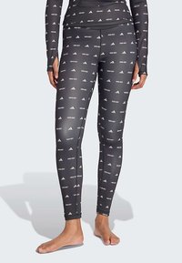 Μαύρες κολάν πλήρους μήκους με λευκό κείμενο "MCMX GET" και λογότυπα της Adidas, φορεμένες από ένα άτομο που στέκεται ξυπόλητο σε ανοιχτό φόντο.