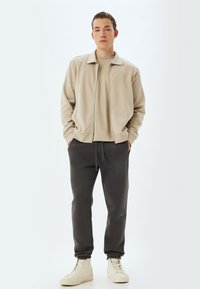Beige zip-upjack met een bijpassende trui, gecombineerd met donkergrijze sweatpants en lichtgekleurde sneakers. Glad materiaal, ontspannen pasvorm.