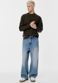 Pulover verde închis cu model în relief, blugi decolorati de culoare albastru deschis, croi lejer, adidași albi cu talpă bej, stând în fața unui fundal simplu.