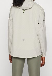 Persona vistiendo una chaqueta con capucha de color beige claro con un pequeño logo de montaña en la parte posterior, combinada con leggings de color verde salvia, de espaldas a la cámara.