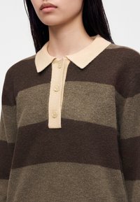 Jeune personne portant un pull en maille rayé marron et olive avec un col crème et trois boutons crème sur le devant.