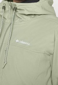 Lichtgroene gewatteerde jas met capuchon, voorzien van een voorkant rits en koordjes, met een zichtbaar Columbia-logo op de borst.