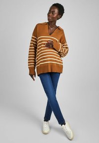 Pull en tricot rayé brun à col, coupe oversized et manches longues, porté avec un jean bleu et des baskets blanches.