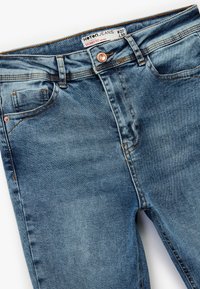 Jeans ajustados de color azul claro fabricados en denim, con un cierre de botón, cinco bolsillos y detalles de costuras en naranja contrastante.