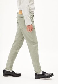 Pantalons en coton vert clair au fit slim, avec des poches latérales et une patch en cuir à l'arrière. Stylés avec des mocassins noirs et une chemise blanche.