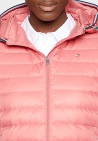 Helle rosa Puffjacke mit horizontalen Nähten, Reißverschluss und Kapuze. Darunter ist ein weißes Hemd und ein kleines Logo-Highlight.