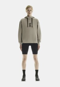 Felpa beige con logo, cordini neri, abbinata a pantaloni corti neri, calze nere e scarpe sportive. Tessuto liscio e vestibilità rilassata.