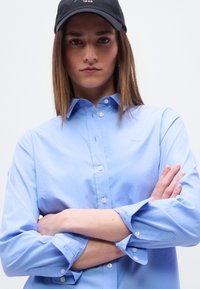 Νεαρή γυναίκα με μαύρο καπέλο και γαλάζιο πουκάμισο με κουμπιά, σταυρωμένα χέρια, κοιτάζει με αυτοπεποίθηση την κάμερα.