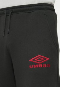 Crne trenirke s vezicom, sa bočnim džepovima i crvenim izvezenim Umbro logom na butini, nosi se uz bijelu majicu.
